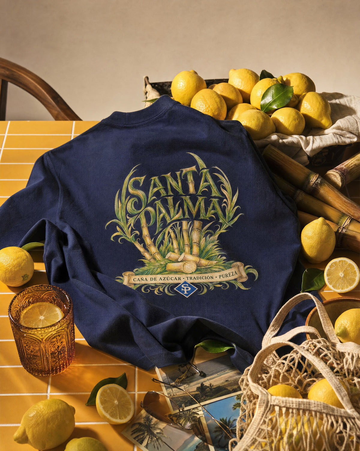 The navy tee spread across a yellow tiled table, back print facing up, with lemons, cut cane, a glass of iced lemonade and old postcards.