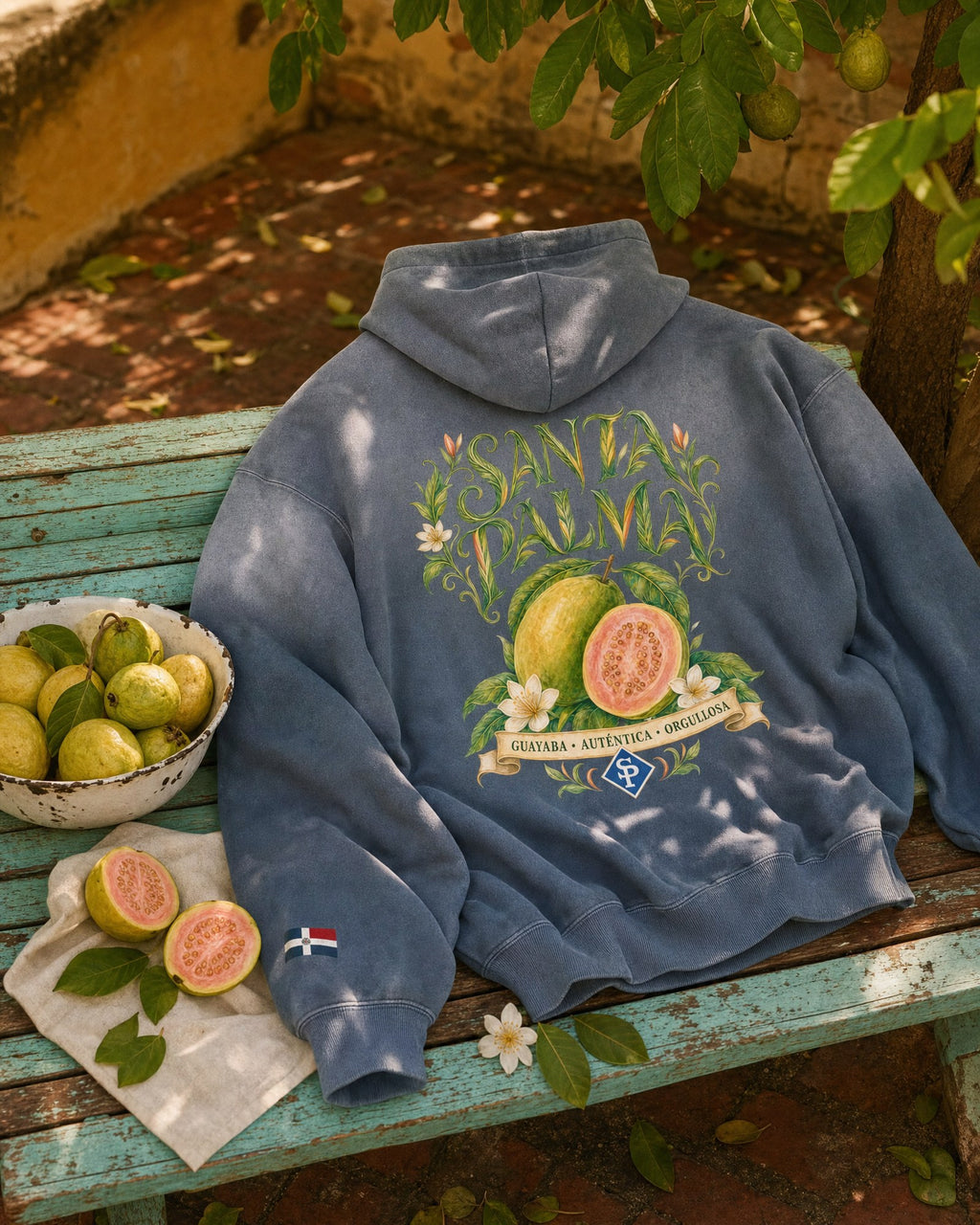 The hoodie laid back-up across a turquoise bench under a guava tree, an enamel bowl of whole guavas beside it and two halved on a cloth.
