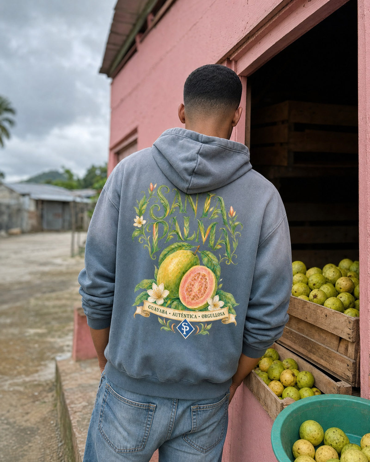 A man from behind in the slate blue hoodie, leaning in the doorway of a pink colmado, crates of guavas at his side under a grey sky.