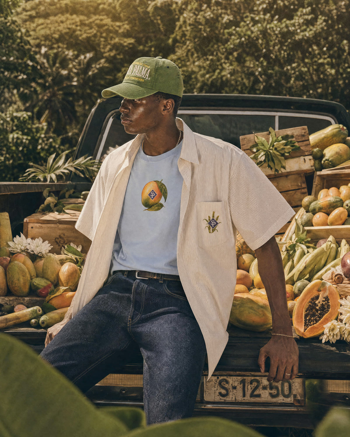 The light blue tee with its mango chest print, worn under an open striped shirt, seated on the tailgate of a pickup loaded with fruit.