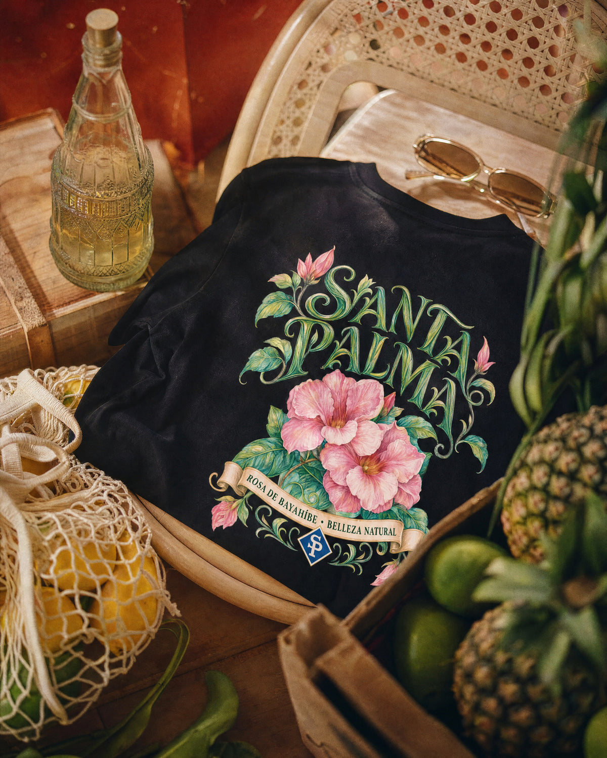 The black tee folded on a caned chair, back print facing up: pink Rosa de Bayahíbe blooms under SANTA PALMA lettering, with pineapples, limes and a net bag of lemons around it.