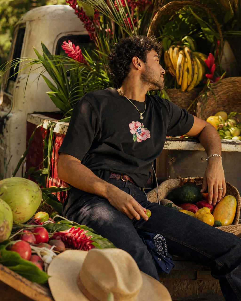 The black tee worn with indigo jeans, its pink Rosa de Bayahíbe chest print in the light, leaning back on a truck bed packed with flowers and fruit.