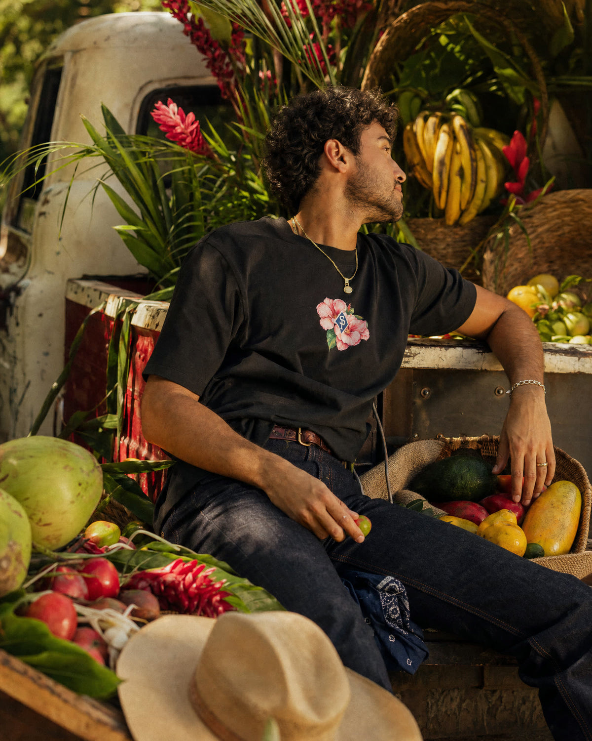 The black tee worn with indigo jeans, its pink Rosa de Bayahíbe chest print in the light, leaning back on a truck bed packed with flowers and fruit.