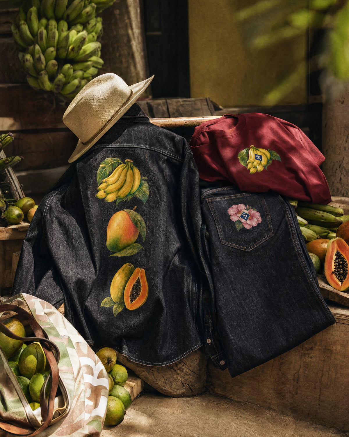 The overshirt spread open over a market block with a straw sombrero at the collar, its back print facing out, folded jeans and a wine guineo tee alongside, limes and cut lechosa banked around it.