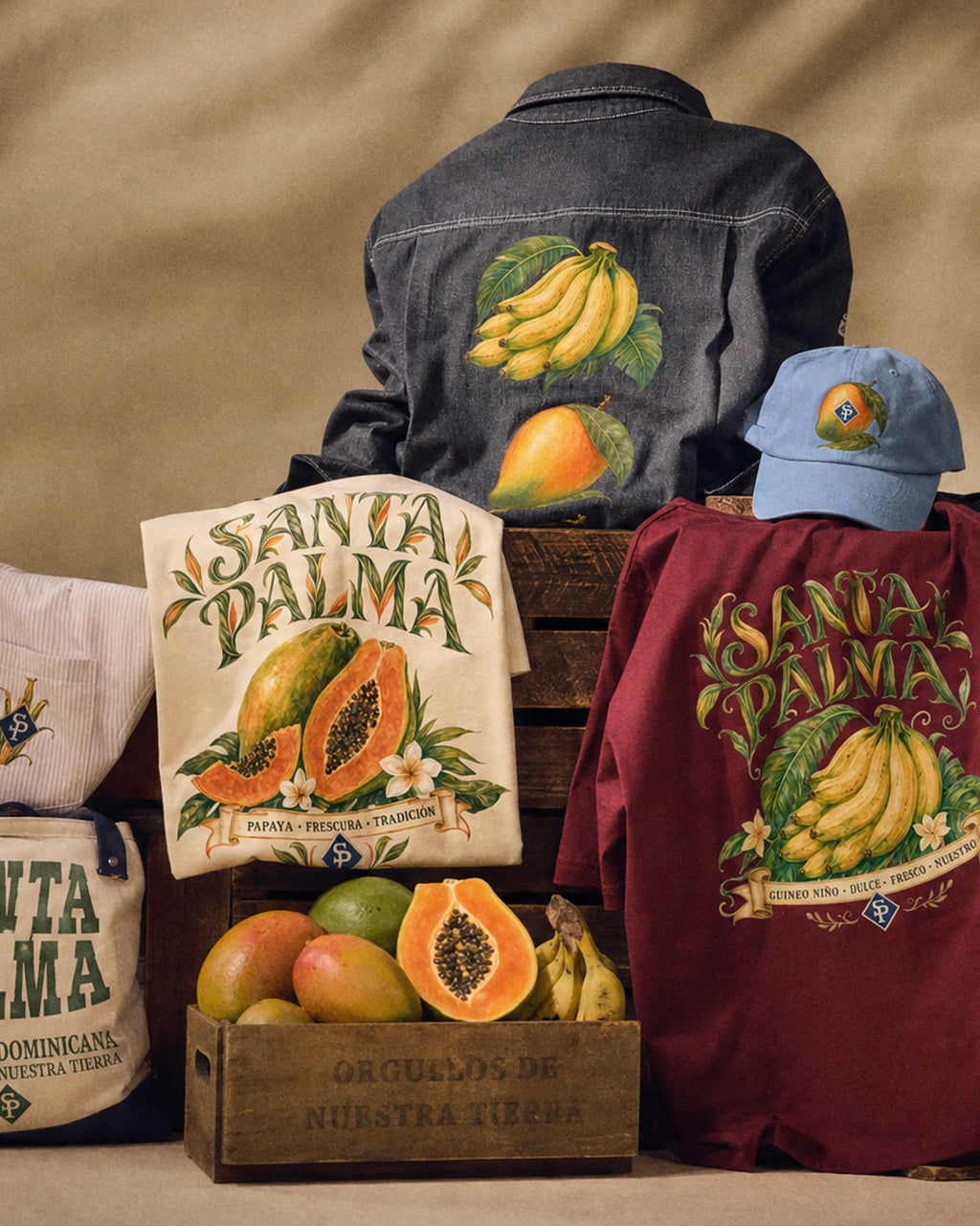 The black overshirt propped upright with its back print showing, guineos above a mango, over a cream papaya tee, a wine guineo tee and a crate stencilled ORGULLOS DE NUESTRA TIERRA.