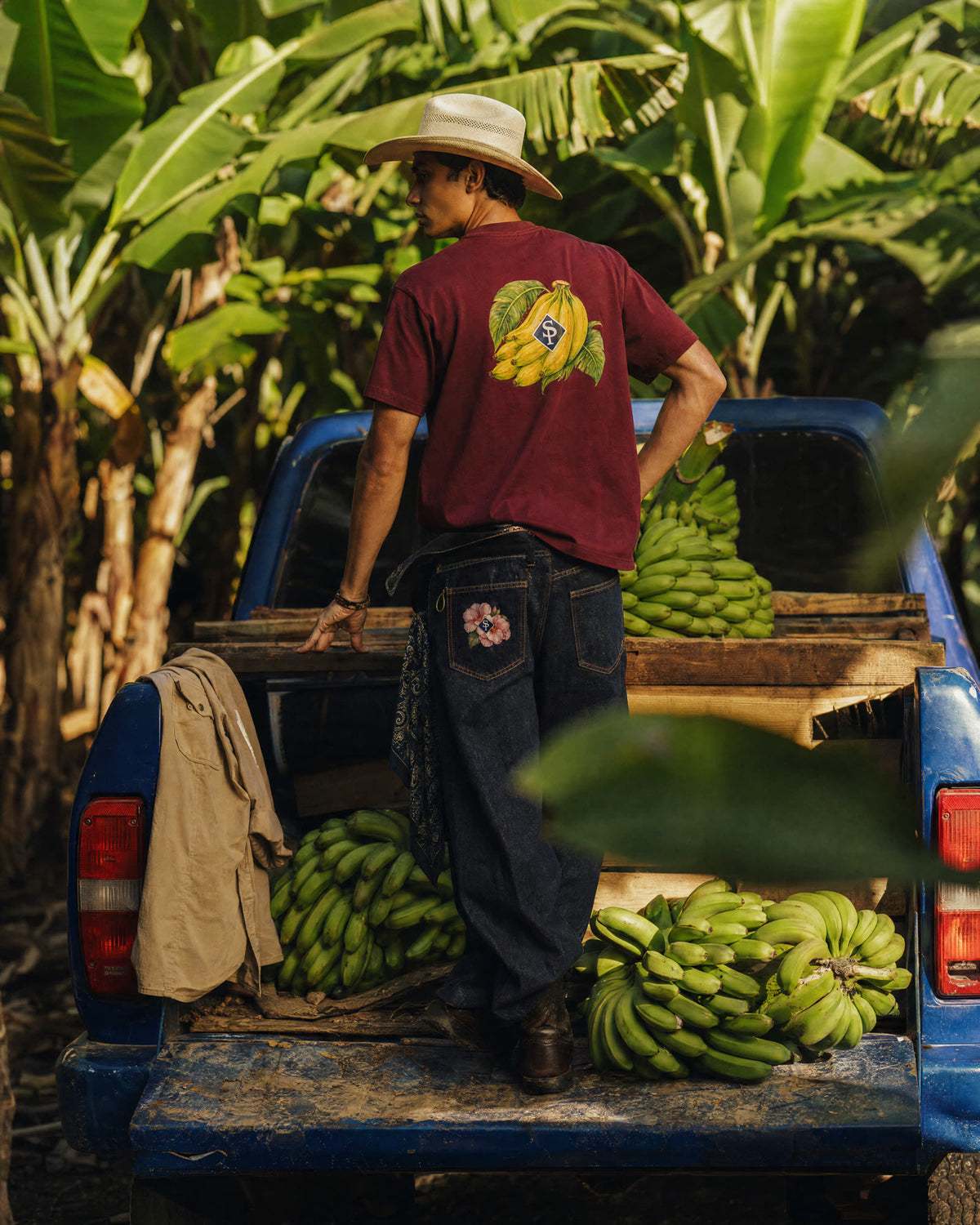 The indigo jeans worn from behind, the Rosa de Bayahíbe embroidery on the back pocket, standing on the bed of a blue pickup loaded with green bananas.